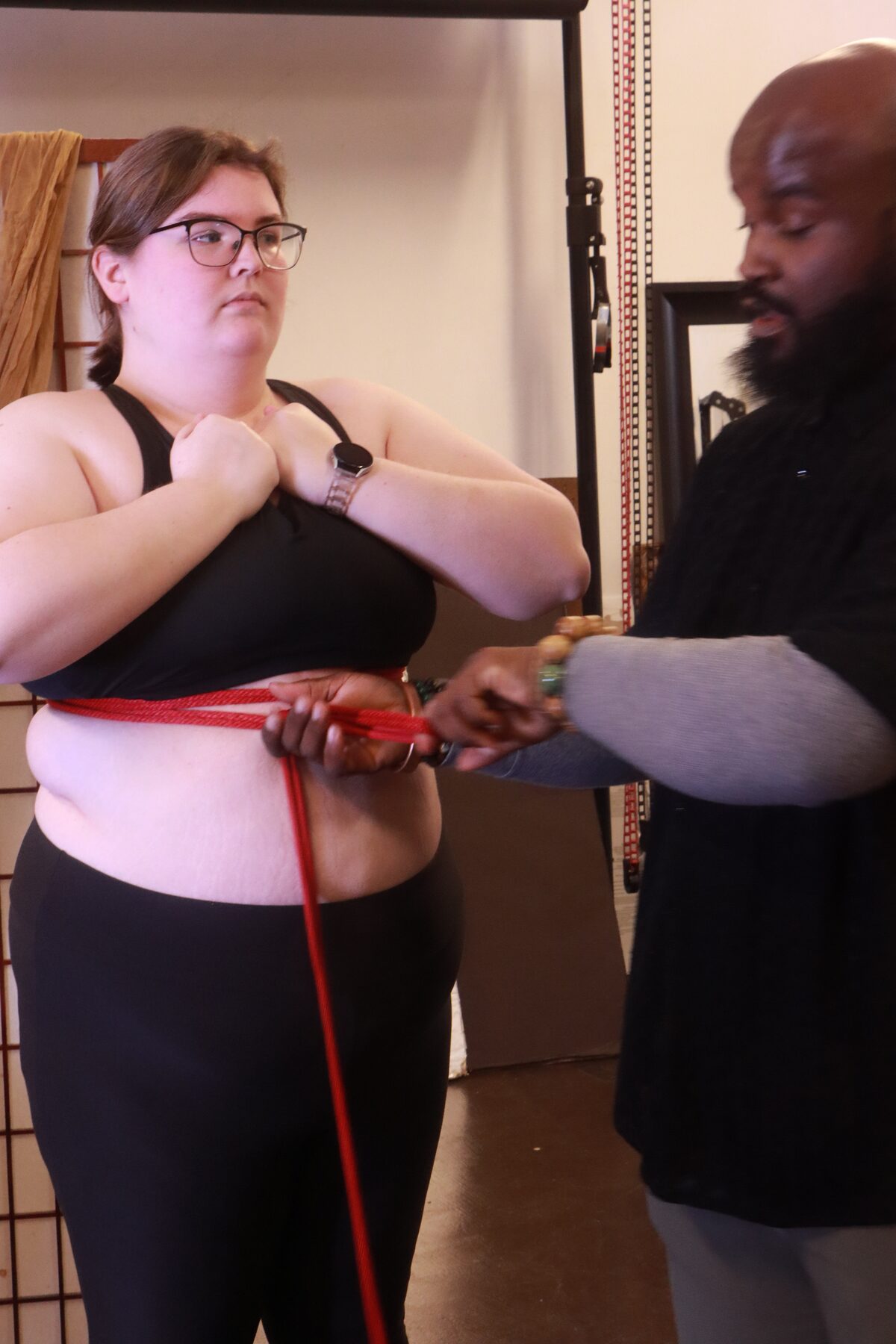 Craig's hands working red rope across a seated student's chest, a close instructional moment
